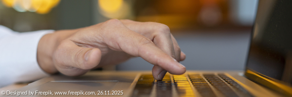 Die Hand eines Mannes bewegt sich auf der Tastatur eines Laptops. Im Hintergrund sieht man veschwommen ein Fesnetr in dem sich Lichter spiegeln 